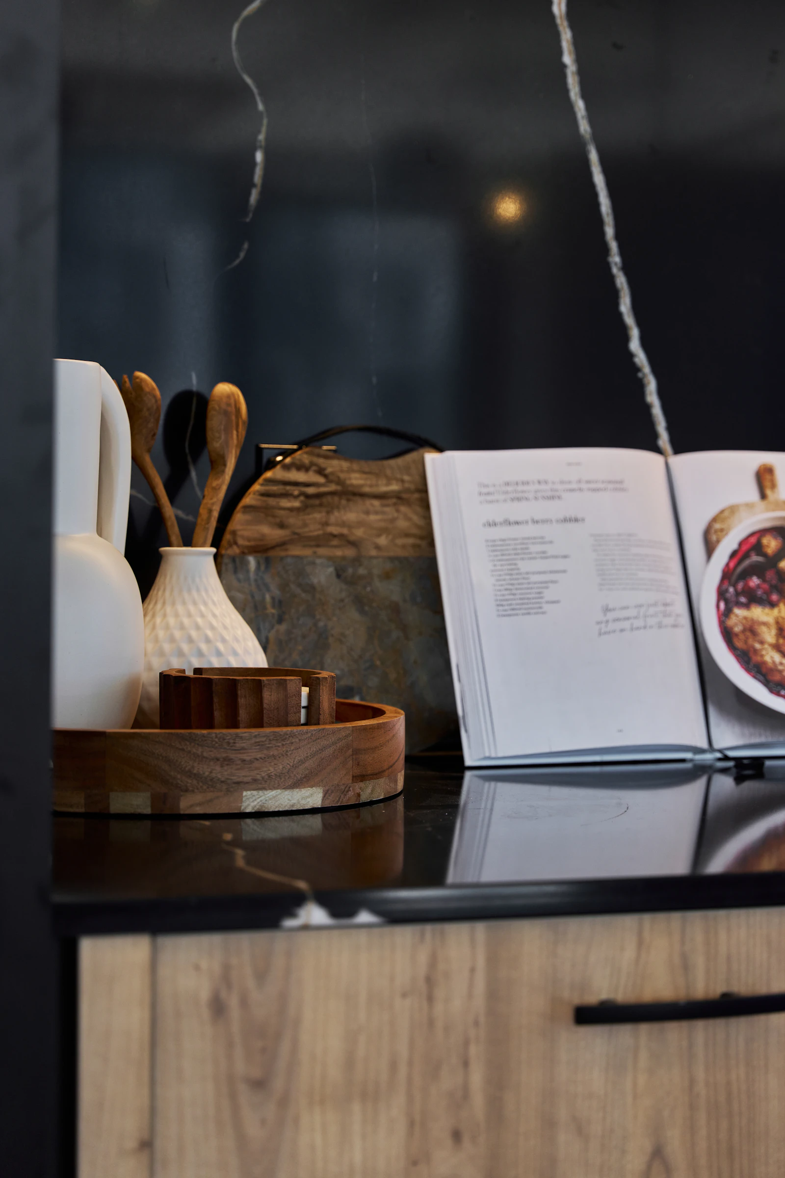 Wooden tray and ceramic utensils beside an open cookbook on a polished dark kitchen counter
